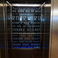 Stationary text on the wall behind the elevators seen through glass. Read fast as you ride!